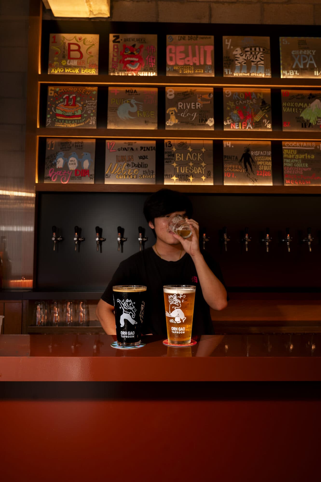 A staff member drinking behind the Orh Gao Taproom counter, with beer taps and illustrated beer boards on the wall behind