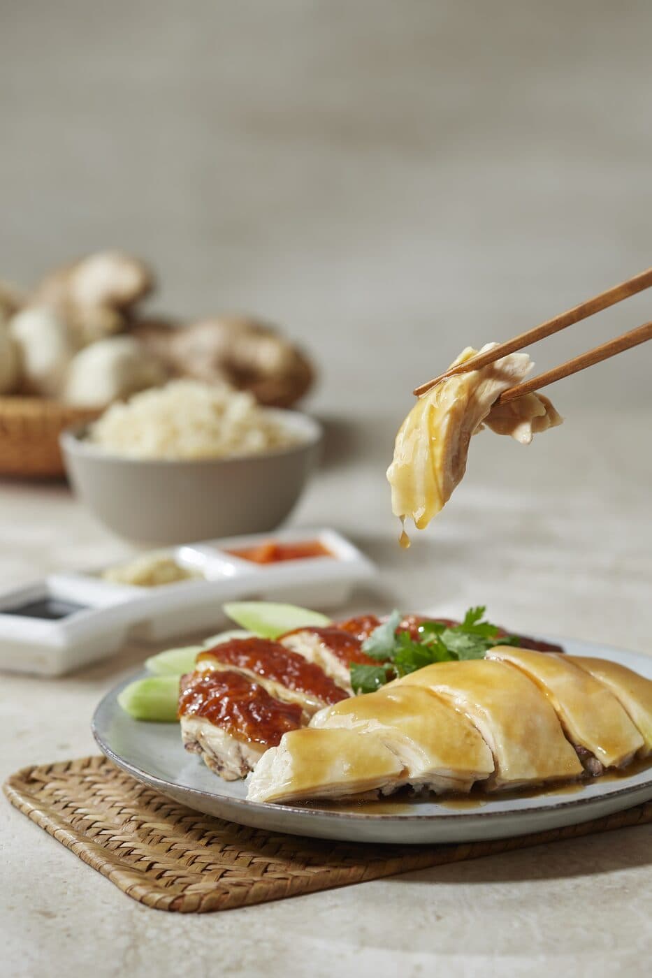 Chopsticks lifting a slice of poached chicken above a plate of chicken with cucumber and sauces