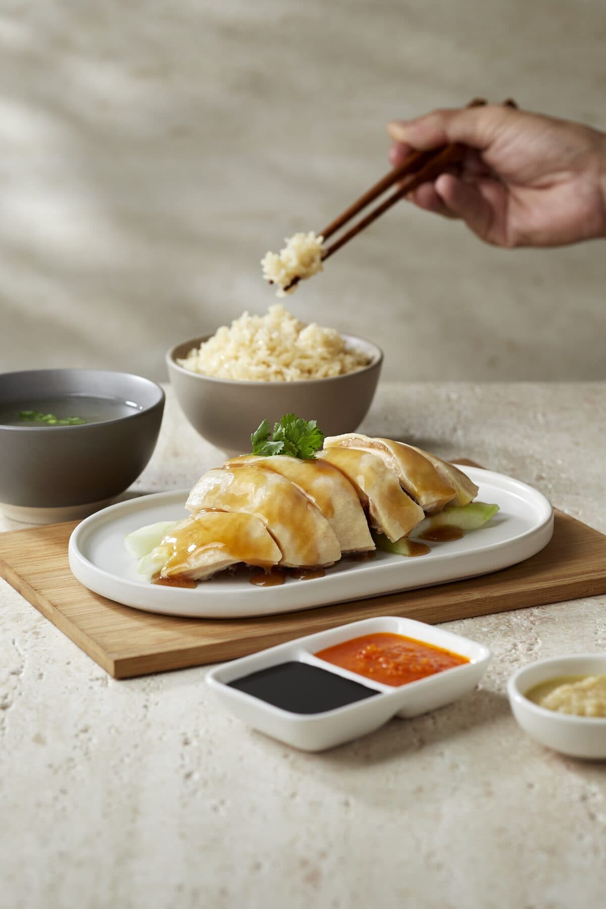 Plate of poached Hainanese chicken with a bowl of chicken rice, soup and chilli and dark soy dipping sauces