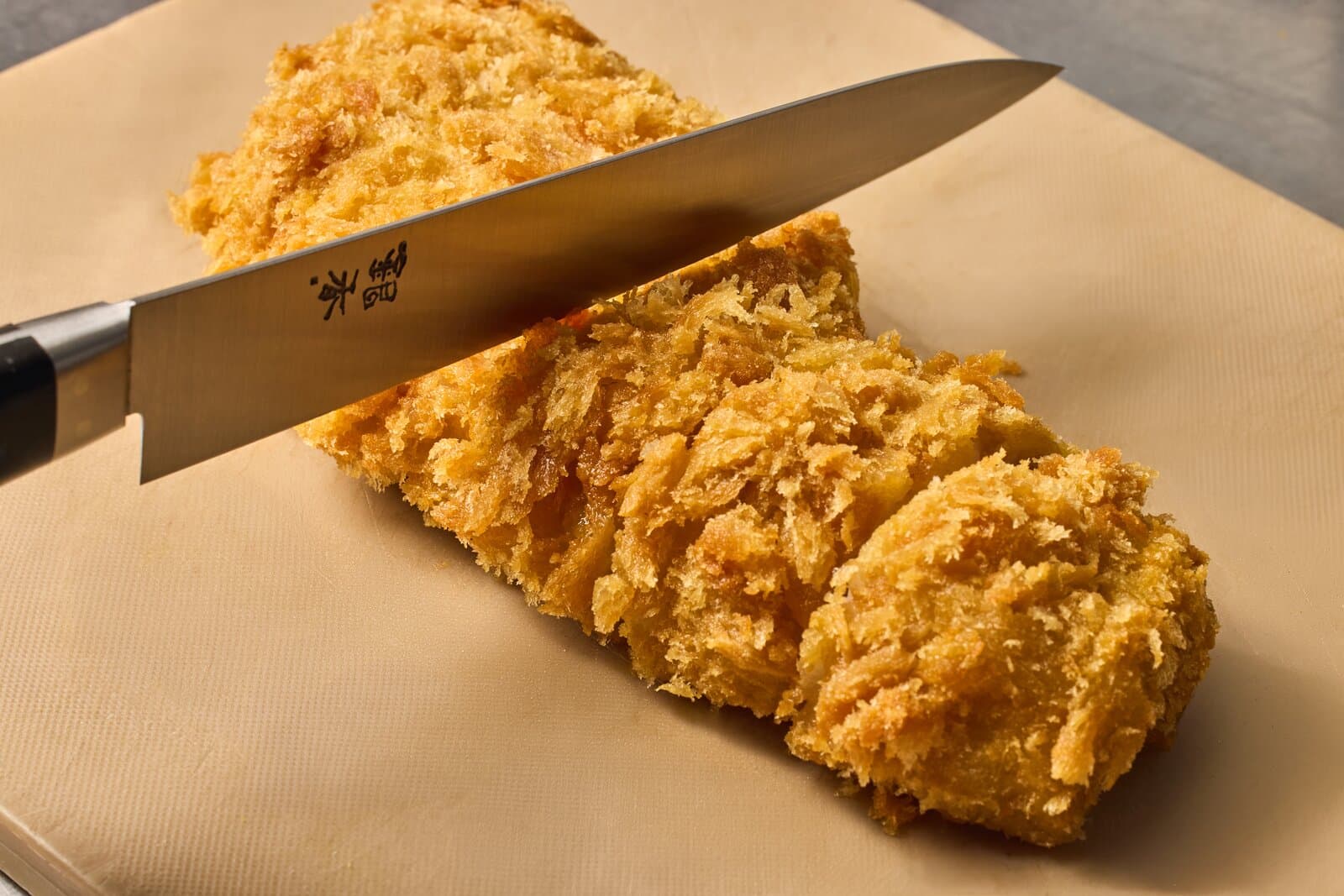 A breaded, deep-fried cutlet sliced into thick pieces on a pale cutting board, with a Japanese chef knife resting across it
