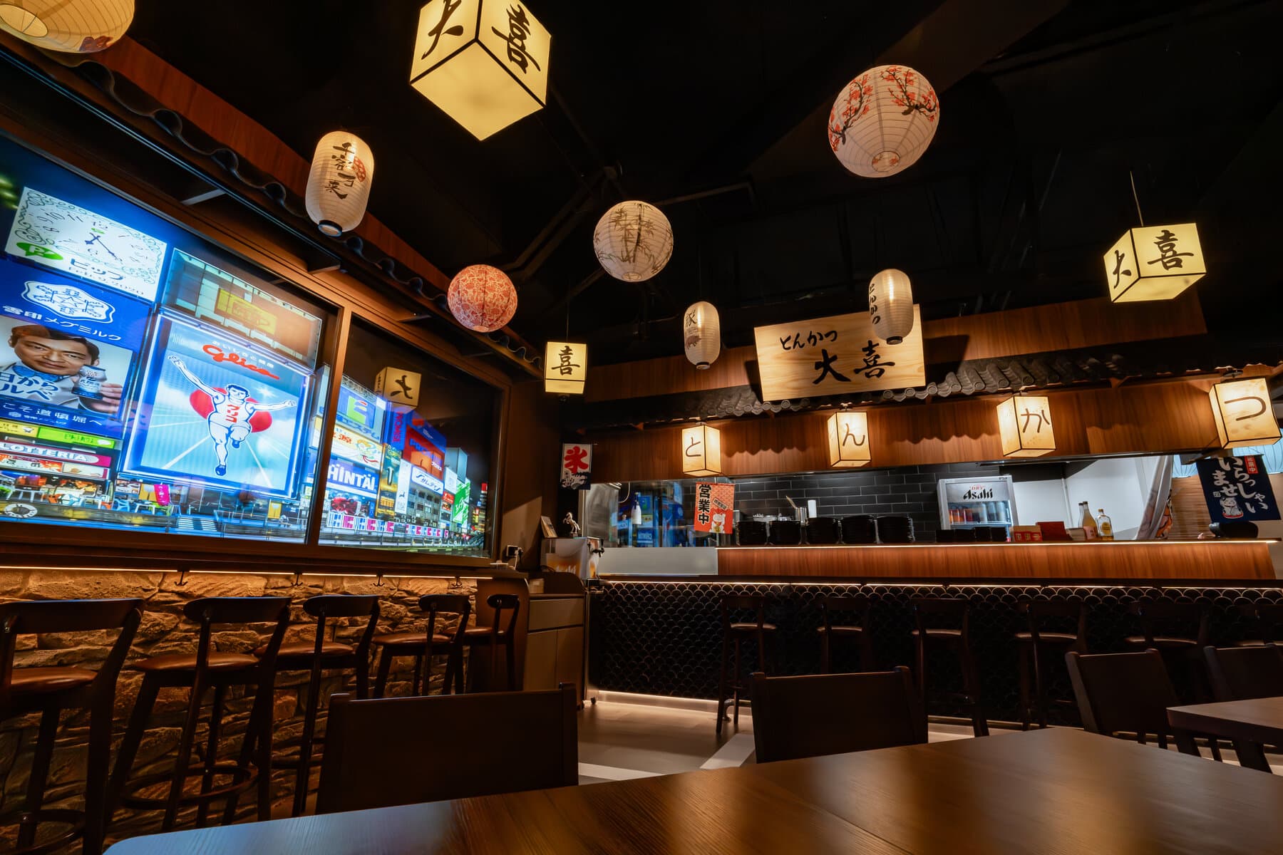 The Tonkatsu Daiki dining room, with printed paper lanterns hung above a long counter of high stools, an illuminated Japanese-script sign on the back wall and a backlit street-scene display beside the counter