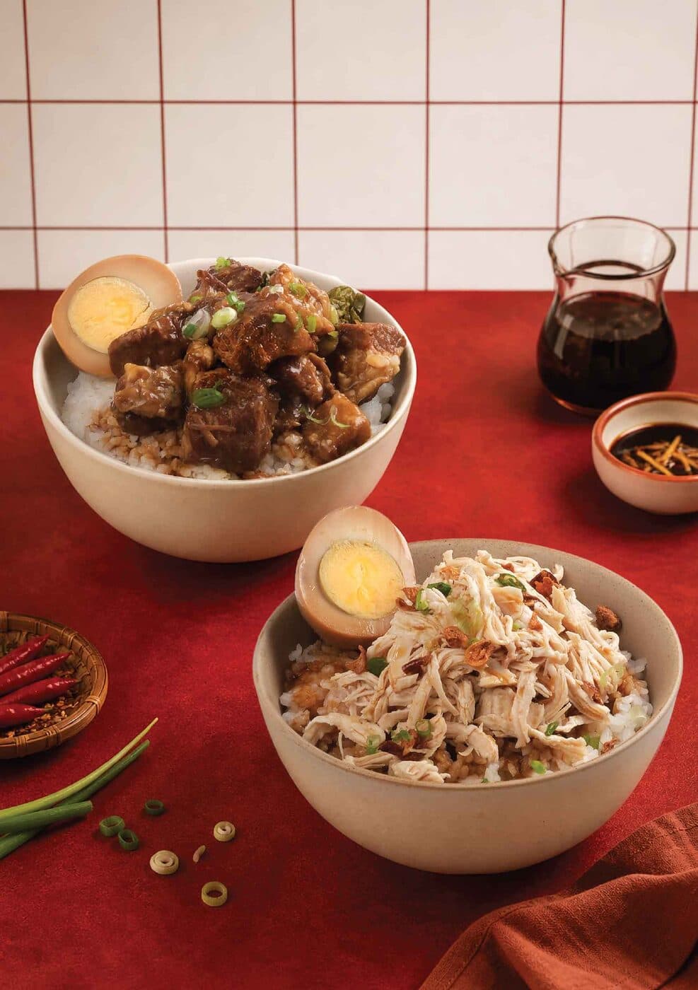 Bowls of braised meat rice and noodles with side dishes on a red surface
