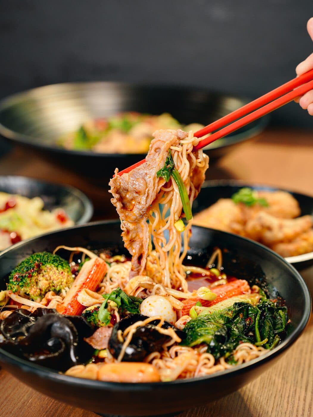 Chopsticks lifting noodles from a mala bowl with greens, mushrooms and sliced meat