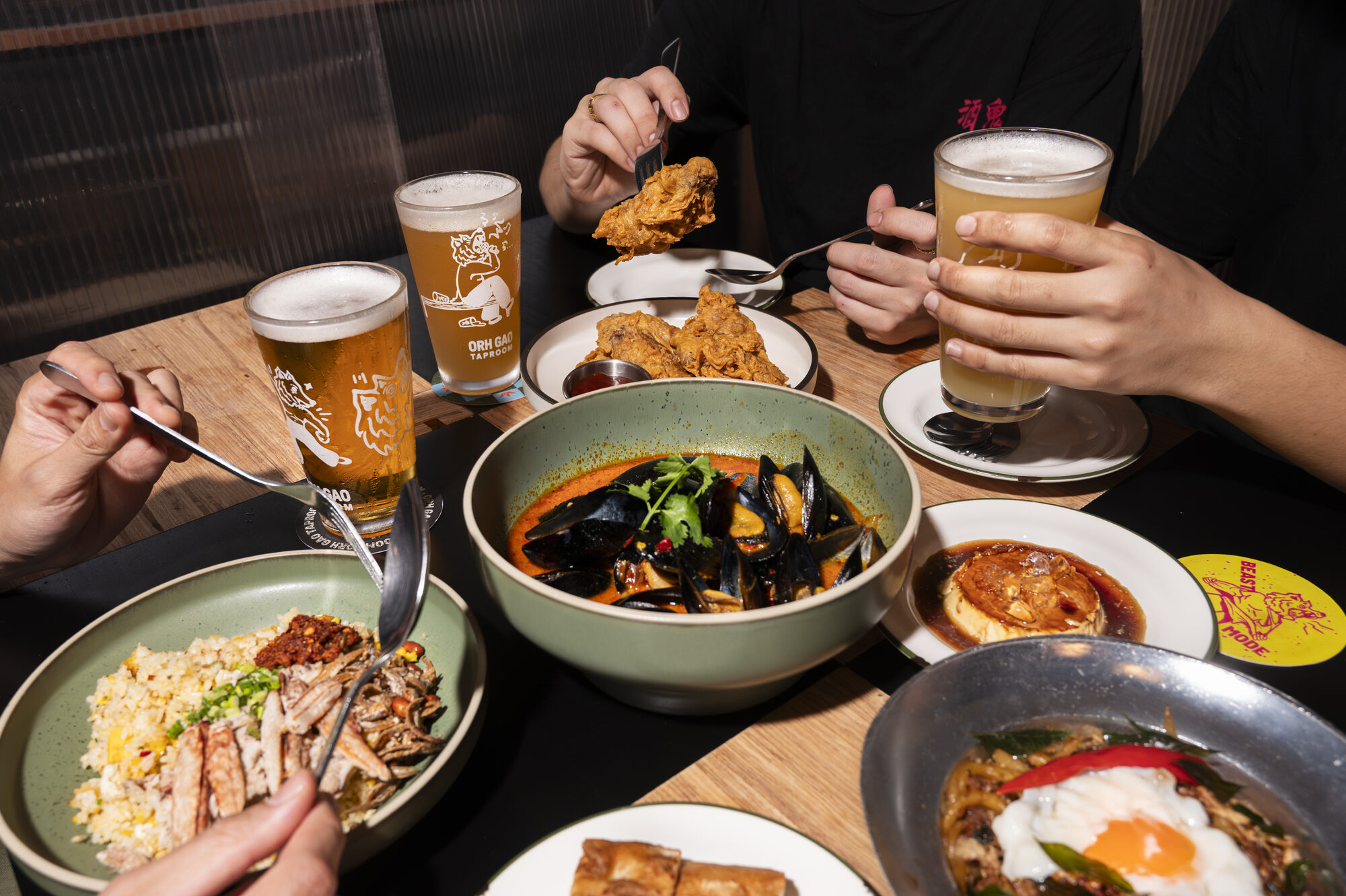Diners serving themselves from shared bowls of noodles, curry and rice at a table set with glasses of beer