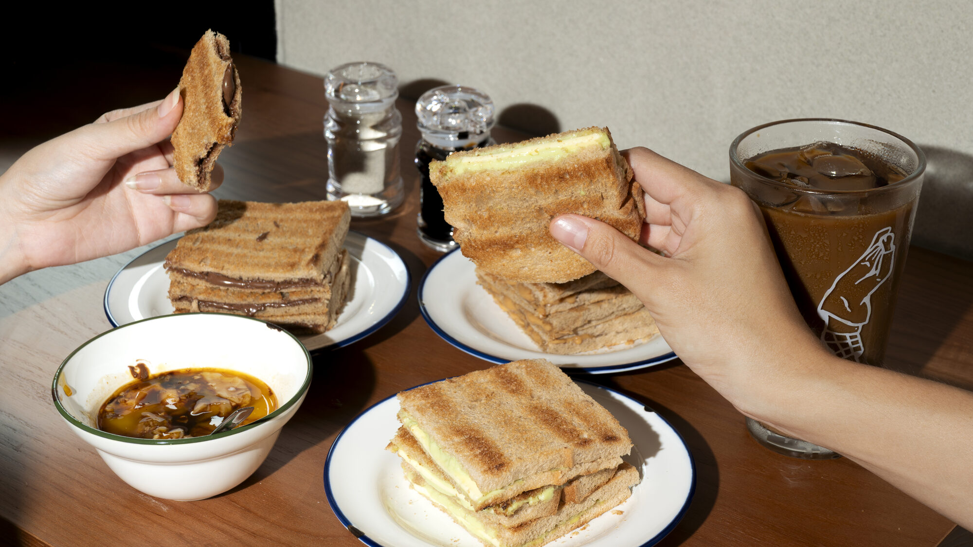 Hands lifting a slice of toast above a bowl of curry, with an iced coffee in a branded glass on the table