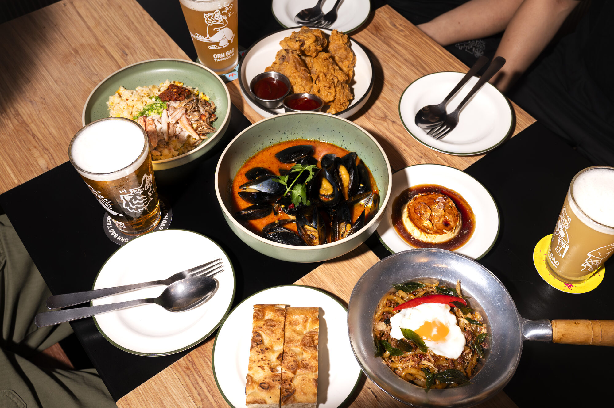 Overhead view of a shared table at Peh Gao Coffeeshop set with fried chicken, a bowl of curry, noodles, toast and a pan of rice topped with a fried egg, alongside glasses of beer