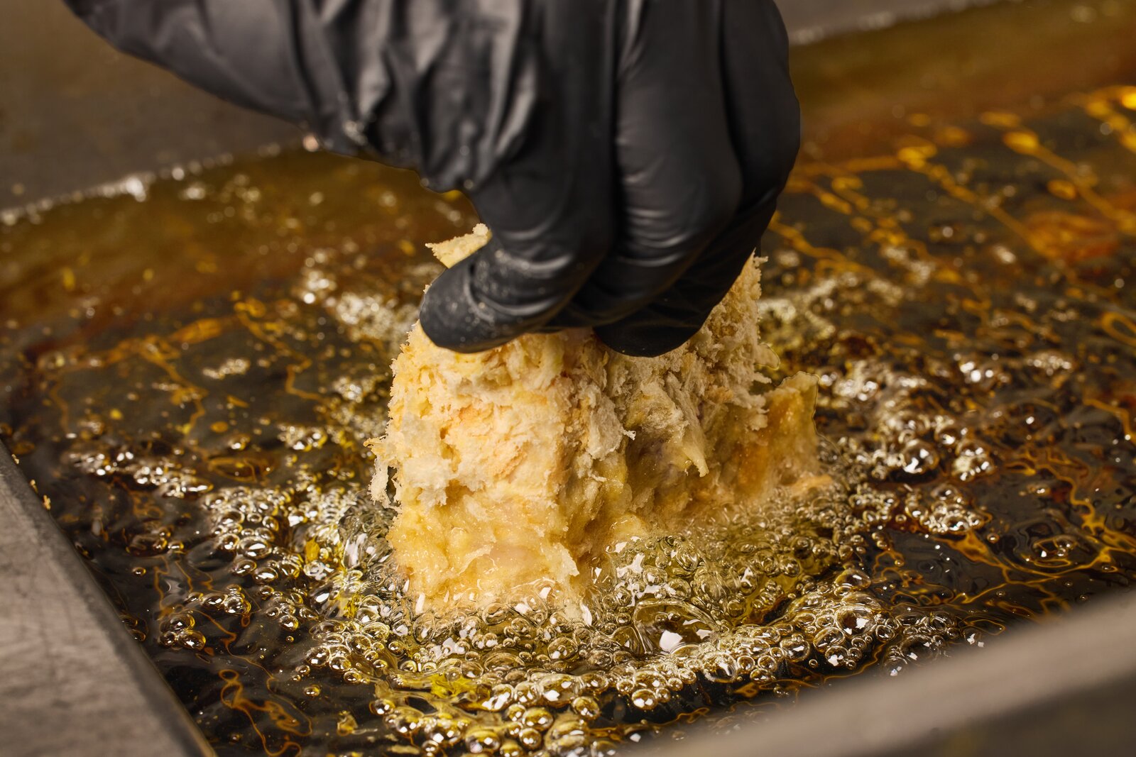 A gloved hand lowering a panko-coated cutlet into a shallow bath of bubbling frying oil